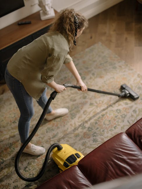 Close-up of a person's hands placing a cleaning cloth into the dust container of a yellow and black vacuum cleaner on a patterned carpet in a room. The vacuum is positioned on the carpet, which appear