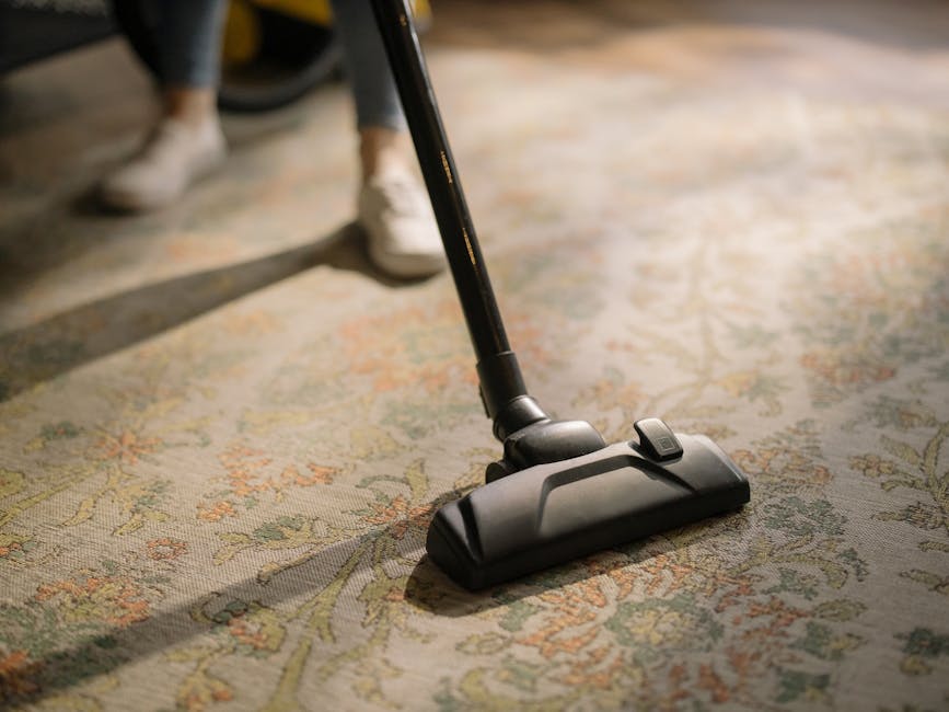 Close-up of a person vacuuming a patterned area rug with a handheld vacuum cleaner, focusing on the dark-colored vacuum head and a section of the floral design on the carpet. The individual is wearing