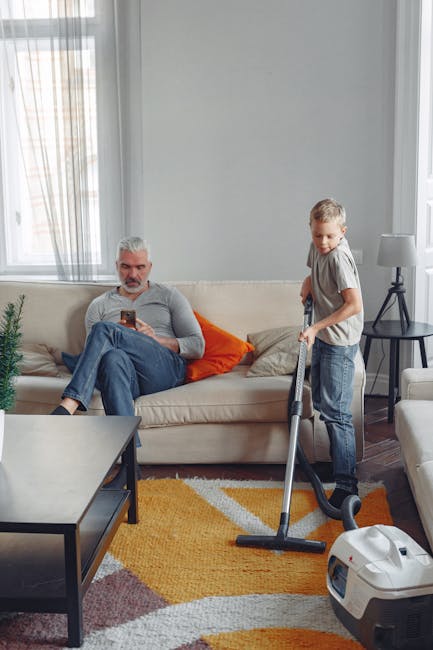 A woman using a yellow vacuum cleaner to deep clean an ornate, beige patterned rug in a living room with wooden flooring and a red leather sofa. She is dressed casually in a beige jacket, jeans, and w